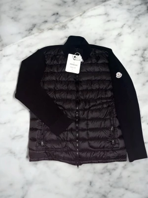 Svart cardiganjacka från Moncler - Snygg svart cardiganjacka från Moncler med quiltad front och stickade ärmar. Jackan har hög krage, dragkedja framtill och två fickor med dragkedja. Moncler-logga på ena ärmen ger en exklusiv touch. Perfekt för dig som vill ha både stil och komfort.