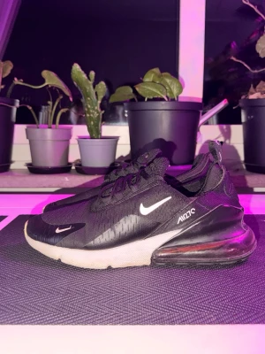 Nike Air Max 270 svarta sneakers - Svarta Nike Air Max 270 sneakers med vit sula och synlig Air-enhet i hälen. Skorna har mesh-överdel för bra ventilation, vita loggor på sidan och tå samt svarta skosnören. Snygg och sportig design som passar till streetwear.