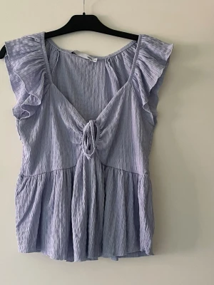 Ljuslila volangtopp med knyt - Supersöt ljuslila topp med volangdetaljer på axlarna och knyt framtill. Tillverkad i ett tunt, krinklat tyg som ger en luftig känsla. Perfekt för varma dagar och ger en romantisk vibe till din outfit.