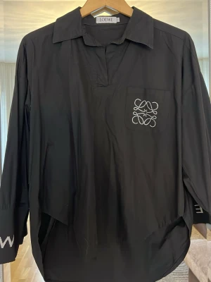 Svart henley-skjorta från Loewe - Svart henley-skjorta från Loewe med klassisk krage och lång ärm. Bröstficka med broderad Loewe-logga i vitt. Skjortan har en avslappnad passform och är tillverkad i mjuk bomull. Perfekt för en stilren och modern look.