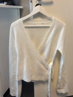 Vit omlottkofta med knytband - Mysig vit omlottkofta med knytband i sidan och djup V-ringning. Koftan har långa ärmar och ribbade kanter. Perfekt för lager på lager och enkel att styla till olika outfits. Aldrig använt för att den inte satt så fint. Men råkade klippa hål där uppe när jag skulle klippa bort lappen. Därav går ner i värde