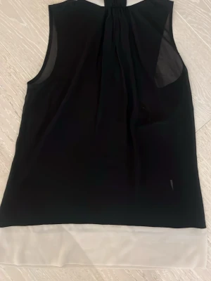 Elegant svart och vit ärmlös blus - Snygg ärmlös blus i svart med vit nederkant. Toppen har en lös passform och lätt transparent material, perfekt för en trendig och stilren look. Passar till både jeans och kjol för en modern outfit. Rök- och djurfritt hem.