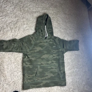 Grön camo hoodie från Hollister - Snygg hoodie i grönt camouflagemönster från Hollister. Klassisk huva med dragsko och stor magficka framtill. Perfekt för en avslappnad streetwear-look. Diskret Hollister-logga på bröstet och mjukt material för extra komfort.