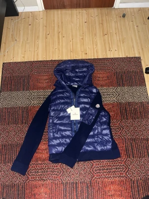 Blå dunväst med huva från Moncler - Snygg blå dunväst från Moncler med stickade ärmar och huva. Västen har en quiltad front i glansigt material, dragkedja och Moncler-logga på ärmen. Perfekt för lager-på-lager och streetstyle. Passar dig som vill ha både värme och stil.