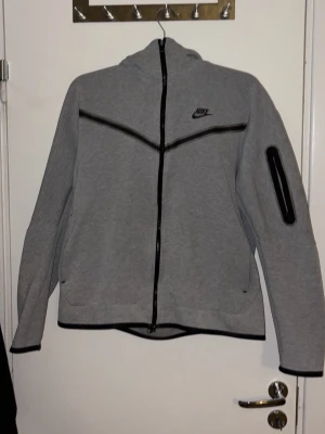 Grå Nike hoodie med dragkedja - Snygg grå hoodie från Nike med hel dragkedja och svarta detaljer. Hoodien har en huva, svarta linjer över bröstet och en praktisk ficka med dragkedja på ärmen. Tillverkad i mjukt sweatshirtmaterial och har Nike-logga på bröstet. L men passar M bra.