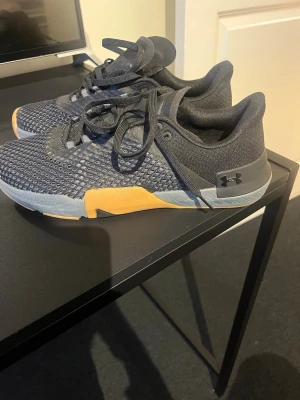 Svarta Under Armour sneakers, aldrig använda - Snygga svarta sneakers från Under Armour med mesh-ovandel och detaljer i grått och gult. Skorna har snörning och låg profil, perfekt för en sportig och avslappnad stil. Sulan är slitstark och ger bra grepp.