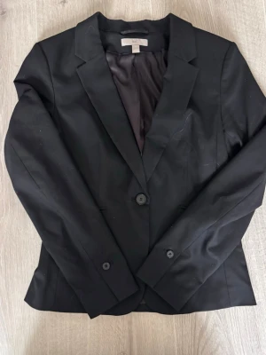 Svart kavaj med två knappar - Stilren svart kavaj med två knappar framtill och klassisk krage. Figursydd modell med helfodrad insida och långa ärmar. Passar perfekt till både skjorta och t-shirt för en snygg look.