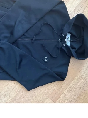 Svart hoodie från Comme des Garçons Play - Säljer en svart hoodie från Comme des Garçons Play med dragkedja framtill och huva med snören. På bröstet sitter det ikoniska svarta hjärtat med ögon. Hoodien är i mjukt bomullsmaterial och har en clean, stilren look.