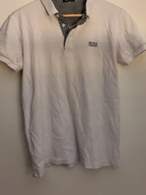 Vit kortärmad t-shirt från BOSS - Vit t-shirt från BOSS med korta ärmar och klassisk krage. Diskret BOSS-logga på bröstet och rutigt tyg på insidan av kragen för en snygg kontrast. Perfekt till jeans eller shorts för en clean look.