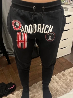 Svarta Hoodrich mjukisbyxor med patchar - Svarta mjukisbyxor från Hoodrich med vita och röda patchar och broderade detaljer framtill. Byxorna har dragsko i midjan och smal passform med muddar vid bensluten. Perfekta för en streetwear-look.