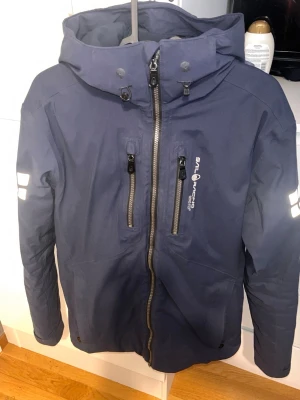 Marinblå vindjacka Sail Racing Gore-Tex - Snygg marinblå vindjacka från Sail Racing med huva, hel dragkedja och två bröstfickor med dragkedja. Jackan har vita logotryck på bröstet, ärmarna och huvan. Tillverkad i ett slitstarkt, vattenavvisande gore-tex material som passar perfekt för blåsiga dagar.