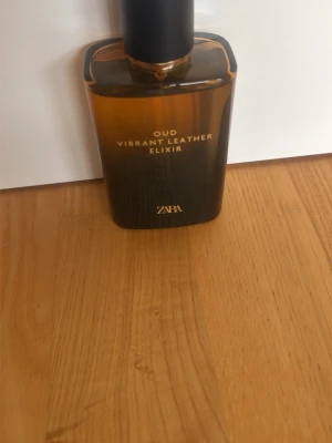 Zara Oud Vibrant Leather Elixir parfym - En elegant parfym från Zara med namnet Oud Vibrant Leather Elixir. Flaskan är fyrkantig och har en snygg bruntonad glasdesign med svart kork. Texten på flaskan är i guld, vilket ger en lyxig känsla. Perfekt för dig som gillar exklusiva dofter.