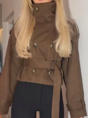 Kort brun trenchcoat från DORA - Snygg kort trenchcoat i mörkbrunt från DORA. Jackan har dubbelknäppning, bred krage, axelklaffar och bälte i midjan. Helt slutsåld med nypris på 1500kr🩵🩵