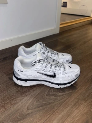 Nike P-6000  - Nike P-6000 sneakers i vitt mesh och syntet med svarta detaljer och silvriga inslag. Klassisk låg modell med svart Swoosh på sidan och coola detaljer på ovandelen. Perfekta för dig som gillar sportig och trendig stil. Skorna har använts varsamt, storlek 38,5.