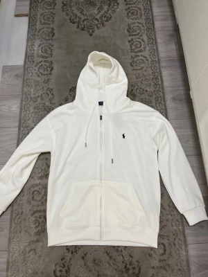 Vit hoodie  Ralph Lauren - Snygg och klassisk broderad logga på bröstet. Hoodien har justerbar huva med snören och en stor känguruficka. Tillverkad i mjukt material som känns skönt mot huden. Perfekt för en clean och stilren look.