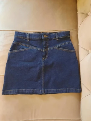 Blå jeanskjol från Molo -  Snygg blå jeanskjol från Molo med klassisk femficksdesign, bälteshällor och knappstängning fram. Kjolen har kontrastsömmar och är i en medellång modell som passar perfekt till många olika stilar. Tillverkad i slitstarkt denimtyg. Är strl 176 man passar som S/M🤍det är en mörkare färg i verkligheten bara så ni vet fast syns inte på kameran. Köptes in för 900 kr men säljer för 500kr😛