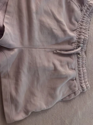 Ljuslila mjukisshorts med dragsko - Säljer ett par ljuslila mjukisshorts med bred resår i midjan och dragsko med metallöljetter. Shortsen har en avslappnad passform och är tillverkade i ett mjukt sweatshirtmaterial. Perfekta för chill dagar eller träning.