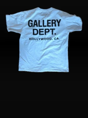 Vit t-shirt från Gallery Dept. - Vit t-shirt från Gallery Dept. med svart tryck både fram och bak. På bröstet står det 'GALLERY DEPT.' och på ryggen 'GALLERY DEPT. HOLLYWOOD, CA.'. Klassisk passform och rund hals. Tillverkad i mjuk bomull för skön känsla.