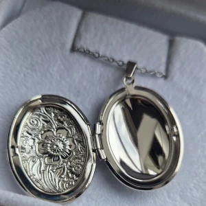 Silverfärgat ovalt medaljonghalsband - Säljer ett silverfärgat halsband med en oval medaljong som har vackra blommönster ingraverade på framsidan. Medaljongen går att öppna och har plats för små bilder eller minnen. Kedjan är tunn i rostfritt stål och stilren, perfekt för dig som gillar detaljerade smycken. Kedjans längd 45 cm och hänget ca 3,4 cm. 