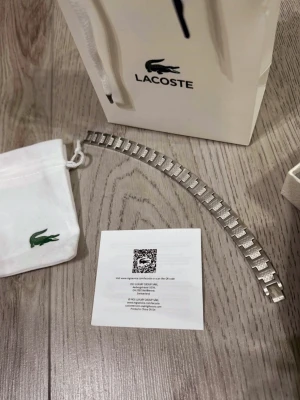 Silverfärgat armband från Lacoste - Snyggt silverfärgat armband från Lacoste med länkdesign och diskret krokodillogga på spännet. Armbandet är tillverkat i metall och har en stilren, modern look som passar perfekt till din outfit. Levereras med originalpåse och presentpåse från Lacoste.