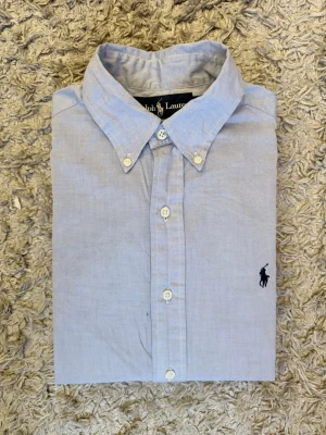 Ljusblå skjorta från Ralph Lauren - Klassisk ljusblå skjorta från Ralph Lauren med button-down krage och broderad mörkblå logga på bröstet. Skjortan har långa ärmar, vita knappar och är tillverkad i ett tunt bomullstyg. Perfekt för en clean och stilren look.