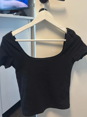 Svart croppad topp med vid urringning - En enkel och stilren svart croppad topp med korta ärmar och bred, djup urringning. Perfekt att matcha med höga jeans eller kjol. Tillverkad i mjukt stretchigt material för en bekväm passform.