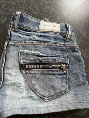 Blå jeansshorts med dragkedjor - Säljer ett par blå jeansshorts med slitna detaljer och coola dragkedjor på bakfickorna. Shortsen har klassisk femficksdesign, bälteshällor och kontrastsömmar. Perfekta för en avslappnad och trendig look.