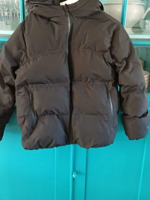 Svart pufferjacka från Zara - Svart pufferjacka från Zara i storlek 164 (13-14 år). Jackan har huva, två stora fickor med dragkedja och en rejäl vaddering som håller dig varm. Perfekt för kalla vinterdagar och har en klassisk, stilren look.