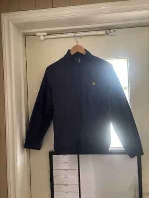 Mörkblå vindjacka från Lyle & Scott - Snygg mörkblå vindjacka från Lyle & Scott med dragkedja framtill och klassisk gul logga på bröstet. Jackan har hög krage, två sidofickor och resår vid ärmslut och midja. Perfekt för blåsiga dagar och enkel att matcha med olika outfits.