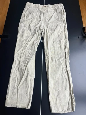 Beiga linnebyxor med rak passform - Snygga beiga linnebyxor med rak passform och elastisk midja baktill. Byxorna har knappgylf och en bakficka med knapp. Tillverkade i ett luftigt linnetyg som passar perfekt till sommaren. Enkla att matcha med olika stilar.