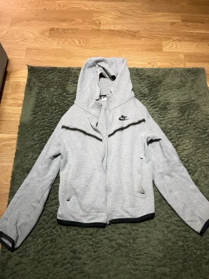 grå Nike tech  - Snygg ljusgrå hoodie från Nike med dragkedja framtill och svart Nike-logga på bröstet. Hoodien har två fickor, svarta detaljer längs dragkedjan och muddar vid ärmslut och nederkant. Perfekt för en avslappnad och sportig stil.
