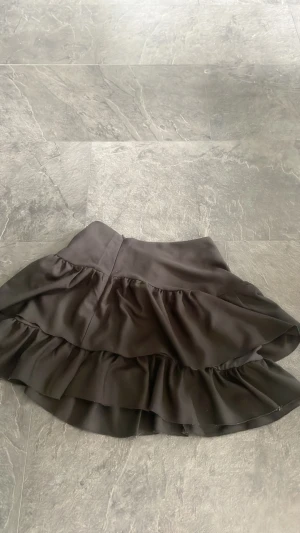 Svart volangkjol med bred midja - Säljer en svart kjol med dubbla volanglager och bred midja. Kjolen har en mjuk, flowy känsla och är perfekt för dig som gillar en feminin och trendig stil. Materialet känns som polyester och kjolen har en snygg, enkel design utan extra detaljer.