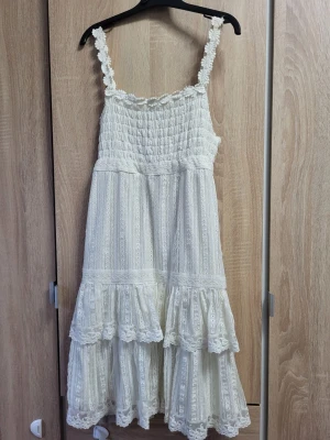Vit spetsklänning med volang - Romantisk vit klänning med smala axelband och vackra spetsdetaljer. Klänningen har volanger nertill och broderade blommor längs banden och midjan. Perfekt för sommarens alla tillfällen och ger en bohemisk känsla.