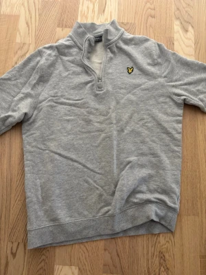 Grå halvzip sweatshirt från Lyle & Scott - Snygg grå sweatshirt från Lyle & Scott med halvzip och ståkrage. Tröjan har det klassiska gula logomärket på bröstet och är tillverkad i mjukt bomullsmaterial. Perfekt för en avslappnad och stilren look.