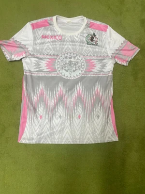 Mexikansk fotbollströja i vitt och rosa - Snygg fotbollströja från Mexikos landslag i vitt, grått och rosa med aztekiskt mönster och tryckt landslagsemblem på bröstet. Tröjan har korta ärmar och rund hals, tillverkad i lätt och ventilerande material perfekt för träning.