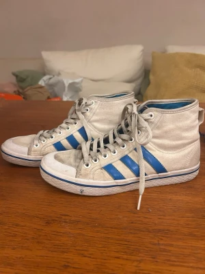 Vita Adidas high top sneakers blå ränder - Säljer ett par vita sneakers från Adidas med klassiska blå ränder på sidorna. Skorna har snörning hela vägen upp och en vit sula med blå detalj. Säger då de inte längre kommer till användning, öppen för att diskutera pris💕