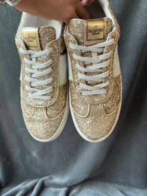 Guldglittriga sneakers från Valentino - Säljer ett par riktigt snygga sneakers från Valentino Garavani med guldglittrande ovandel och vita detaljer. Skorna har vita snören, vit sula och coola nitar bak på hälen. Perfekta för dig som vill sticka ut med stil och älskar glitter.
