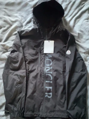 Svart vindjacka från Moncler - Snygg svart vindjacka från Moncler med huva och dragkedja framtill. Jackan har Moncler-logga på ärmen och stor logotyp på bröstet. Tillverkad i lätt, vattenavvisande material och har en bröstficka med dragkedja. Perfekt för blåsiga dagar.
