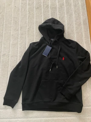Svart hoodie från Polo Ralph Lauren - Svart hoodie från Polo Ralph Lauren med klassisk röd broderad logga på bröstet. Hoodien har huva med dragsko, magficka och ribbade muddar. Tillverkad i mjukt bomullsmaterial, perfekt för en avslappnad och stilren look.
