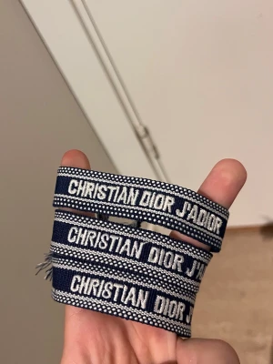Blått armband Christian Dior J'Adior - Snyggt blått tygarmband från Christian Dior med texten 'J'Adior' broderad i vitt. Armbandet har en bohemisk känsla med fransar i ändarna och är justerbart för att passa olika handleder. Perfekt accessoar för att lyfta din outfit.