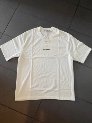 Vit Acne Studios t-shirt i bomull M - Snygg vit t-shirt från Acne Studios med svart broderad logga på bröstet. Klassisk rund hals och korta ärmar. Tillverkad i mjuk bomull för en clean och stilren look. Perfekt basplagg för alla tillfällen.