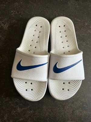 Vita Nike badtofflor med blå logga - Säljer ett par vita badtofflor från Nike med blå swoosh-logga på ovandelen. Tofflorna är i ANVÄNT SKICK.  passar storlek 41
