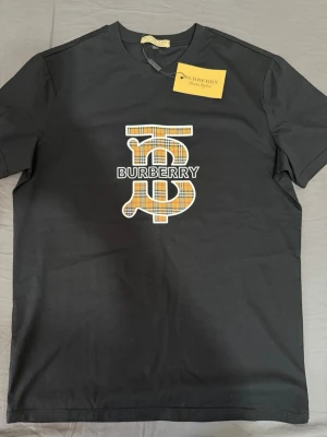 Svart Burberry t-shirt med logga - Snygg svart t-shirt från Burberry med klassisk rutig logga i orange, vit och svart på bröstet. T-shirten har rund hals och korta ärmar. Perfekt för dig som gillar exklusiva märken och stilren design.