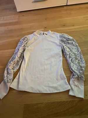 Vit topp med spetsärmar från SHEIN - Trendig vit topp från SHEIN med ribbad kropp och långa, transparenta spetsärmar. Hög halsringning och snygg passform som ger en elegant vibe. Perfekt för dig som vill ha något extra till outfiten.