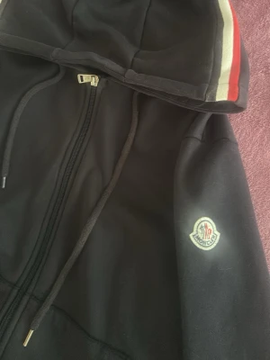 Svart hoodie från Moncler med dragkedja - Snygg svart hoodie från Moncler med dragkedja framtill och huva med vita och röda ränder. Klassisk Moncler-logga på ärmen och fickor på magen. Perfekt för dig som gillar streetwear och vill ha något stilrent med exklusiv känsla.