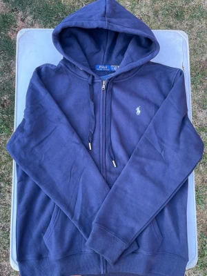 Marinblå hoodie från Polo Ralph Lauren - Snygg marinblå hoodie från Polo Ralph Lauren med dragkedja, huva med dragsko och klassiska loggan broderad på bröstet. Tillverkad i mjukt bomullsmaterial och har fickor framtill. Perfekt för en chill och stilren look.