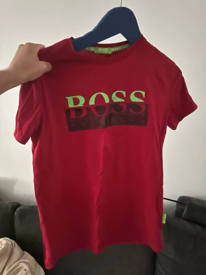 Röd BOSS t-shirt med tryck - Snygg röd t-shirt från BOSS med stort logotryck i grönt och svart på bröstet. Klassisk rund hals och korta ärmar. Perfekt för dig som gillar streetstyle och vill sticka ut med en färgstark look.