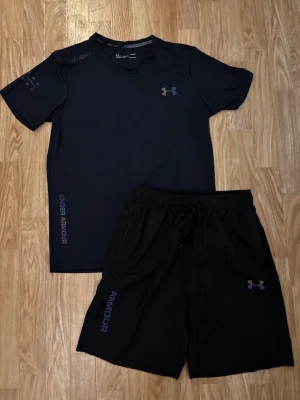 Svart träningsset från Under Armour - Snyggt svart träningsset från Under Armour med t-shirt och shorts. Båda plaggen har logga och textdetaljer i lila och blått. Tillverkade i ett lätt och snabbtorkande material som passar perfekt för gym eller löpning.