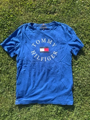 Blå Tommy Hilfiger t-shirt - Snygg blå t-shirt från Tommy Hilfiger med stort tryck och logga på bröstet. Klassisk rund hals och korta ärmar. Tillverkad i mjuk bomull som känns skön mot huden. Perfekt för en avslappnad och stilren look.