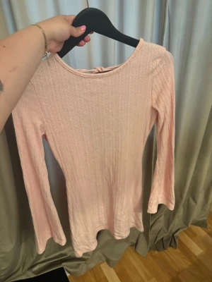 Ljusrosa ribbad topp med öppen rygg - Supersöt ljusrosa långärmad topp med ribbad struktur och öppen rygg med knyt. Perfekt för dig som gillar en feminin och trendig stil. Toppen har en smickrande passform och är tillverkad i ett mjukt material som känns skönt mot huden.